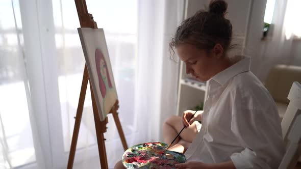Girl Sits on a Chair Near the Easel and Holds a Palette and a Brush in Her Hands alt