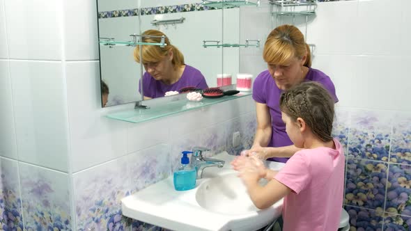 Mom teaches a little girl how to wash their hands with soap over a sink with running water