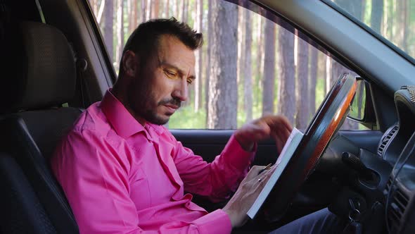 A Sad Businessman in a Car Reads and Writes Notes in a Notebook alt