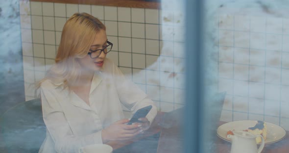 Attractive Young Businesswoman in Glasses Using Smartphone in Cafe View Through the Window alt