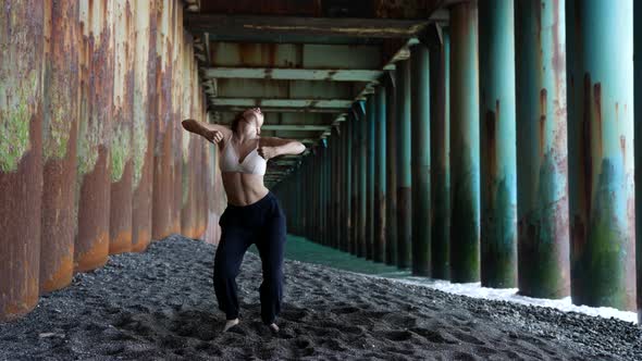 a Barefoot Woman Dances Under the Pillars of the Bridge Against the Background of the Incoming Waves alt