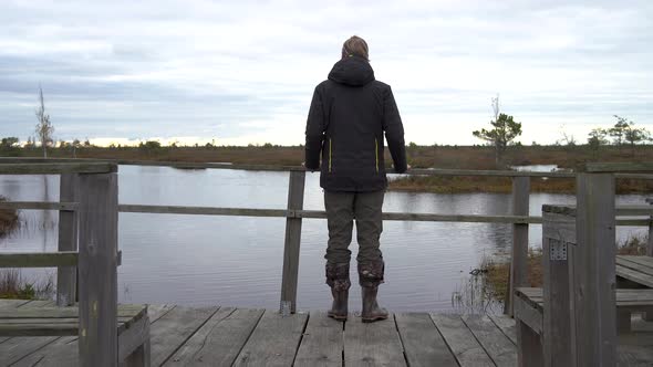Man is Standing on Wooden Bridge Near the Swamp and Looks at Water Back View
