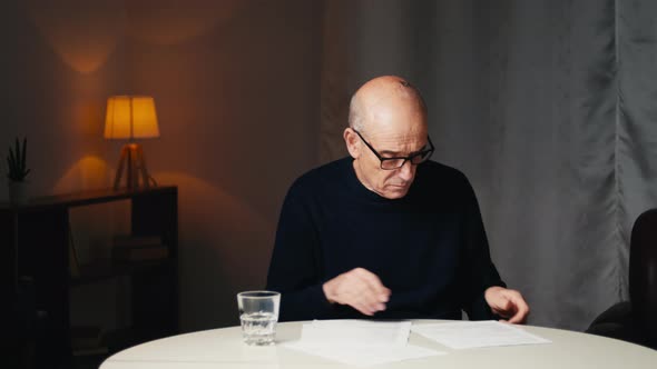Elderly Man Sitting at the Table Reads His Electricity and Gas Bills alt