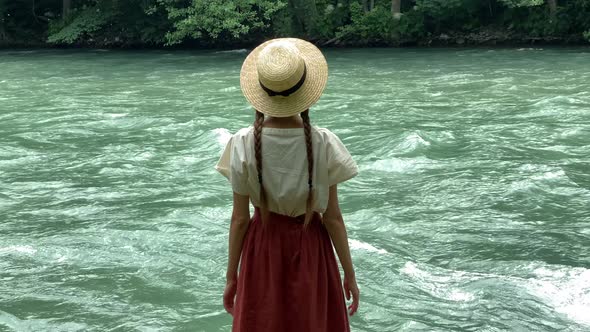 Young vintage woman in dress and straw boater hat stand near rippling mountain river, back view