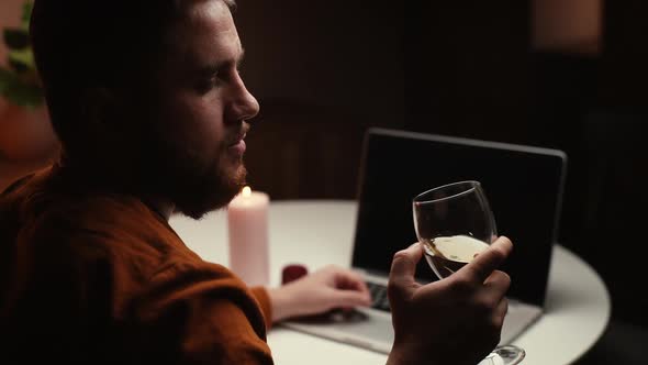 Closeup View of Thoughtful Young Man Drinking Alcohol From Glass During Working on Laptop Computer alt