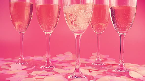Pouring sparkling wine into glasses on a pink background with heart-shaped confetti. Slow motion. alt