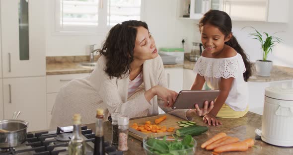 Hispanic mother teaching daughter sitting on countertop cooking, looking at tablet alt