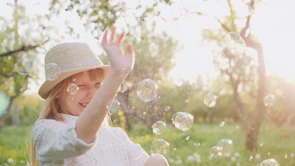 A Cheerful Girl in a Hat Carelessly Plays with Soap Bubbles. Happy Spring and Good Weather alt