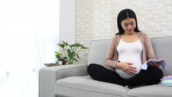 Pregnant young asian woman sitting on sofa with crossed legs touching her belly