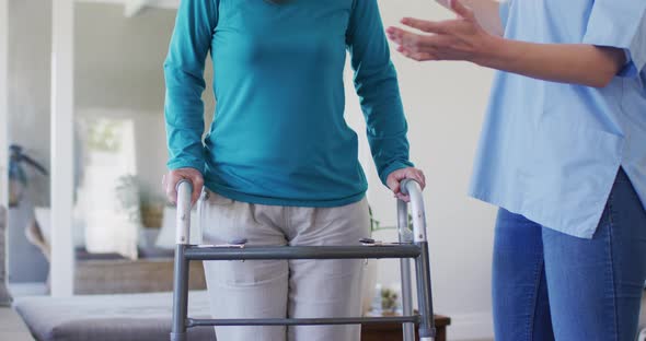 Female health worker assisting senior woman to walk with walking frame at home alt