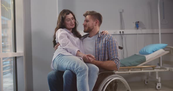 Smiling Woman Sitting on Lap of Disabled Boyfriend in Wheelchair at Hospital