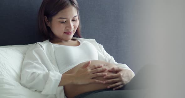 Asian pregnant woman stroking belly while lying on a bed in the bedroom at home.