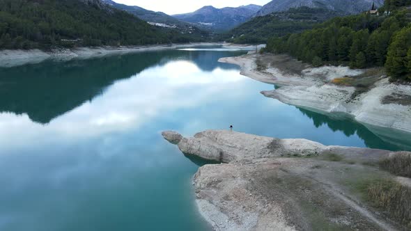 Sinking Aerial of Distant Person By Rocky Shore of Lake in Guadalest alt