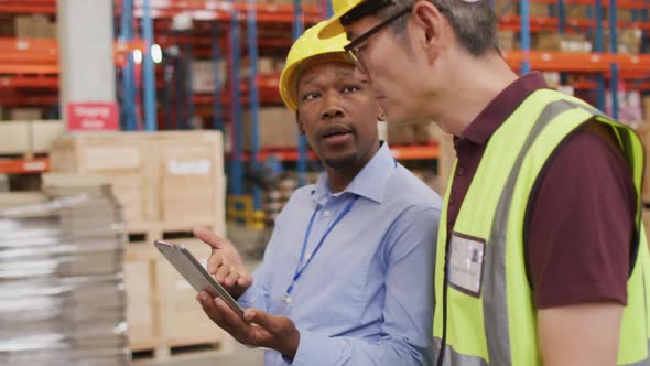 Diverse male workers wearing safety suits and talking in warehouse alt