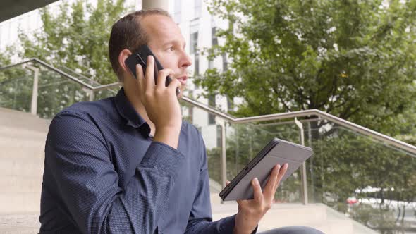 A Caucasian Man Talks on a Smartphone and Holds a Tablet As He Sits on a Staircase in an Urban Area