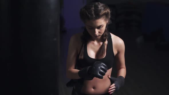 Woman Taking on Boxing Gloves on Hands with Black Boxing Wraps in Dark Room alt