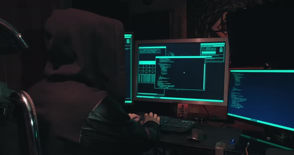Hacker in a Hood Sits in Front of Computer Screens and Hacks Databases alt
