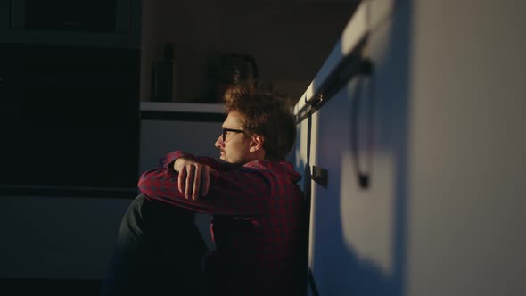 Young man with a thoughtful look sits on the kitchen floor at home and looks ahead alt