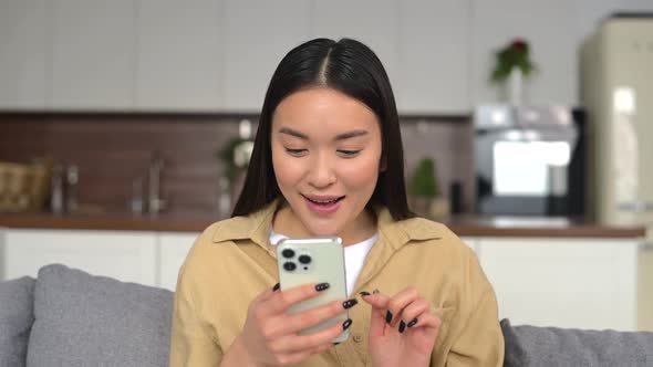 Joyful Asian Woman Using Smartphone Sitting on the Comfortable Sofa at Home