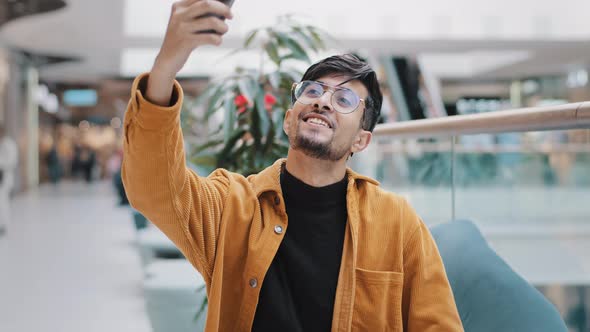 Young Happy Indian Guy Blogger Holds Smartphone Recording Vlog Filming Video for Blog in Mall Joyful alt