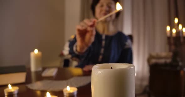 Big White Candle Standing at the Foreground As Blurred Old Caucasian Woman Lighting Up Candles alt