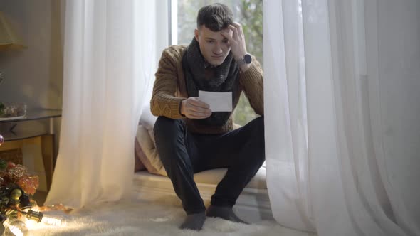 Wide Shot of Depressed Young Man Sitting on Windowsill Looking at Ultrasound Picture alt