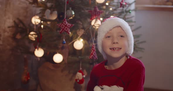 Happy Kid in Santa Claus Hat Posing at Home By Christmas Tree Glowing Lights Slow Motion