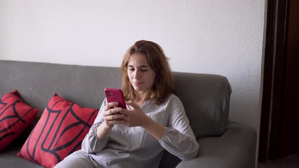 Young Woman Sitting on the Sofa in Pajamas Interacting with Mobile Phone