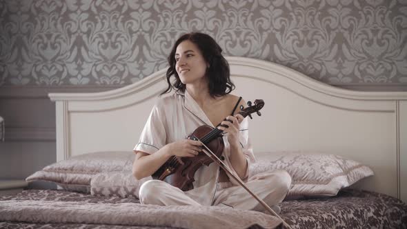 Beautiful Girl Cheerfully Plays the Violin While Sitting on the Bed at Home