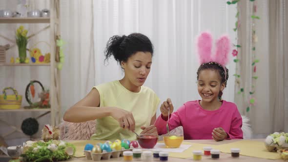 Mom and Daughter with Funny Bunny Ears Dips Eggs in Food Coloring Using Yellow and Red Paint alt