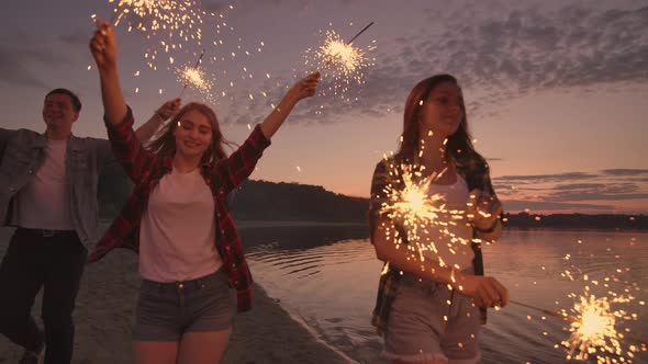 Cheerful Male and Female Friends are Running Along the Beach at Sunset Holding Sparkling Fireworks alt