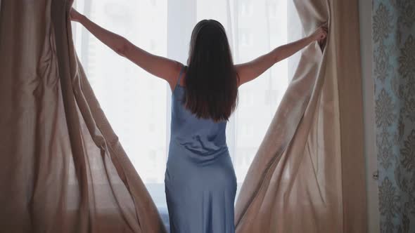 Young Woman Approaches Window Pushes Aside Curtains and White Tulle with Hands Opens Window and