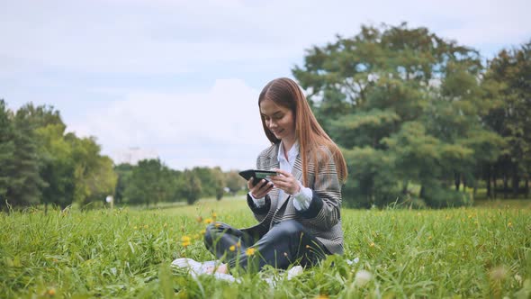 A Young Girl Makes a Purchase with Her Phone and Bank Card in the Park Sitting on the Grass alt