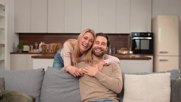 Cheerful Caucasian Couple in Love Sitting on the Sofa in Embrace and Looking at the Camera alt