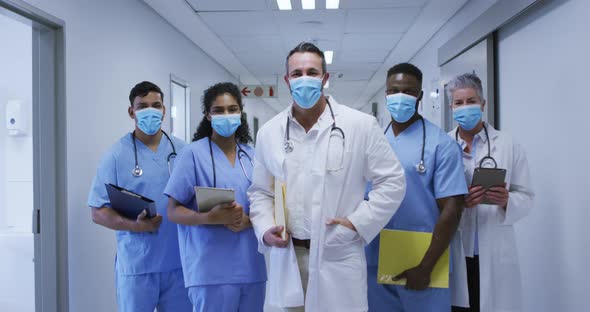 Portrait of diverse group of male and female doctors in face masks standing in hospital corridor alt