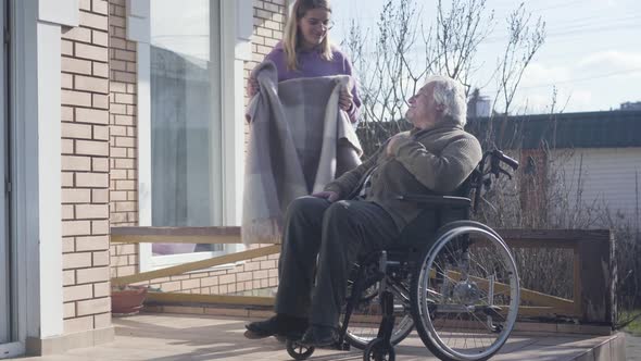 Nice Caucasian Woman in Eyeglasses Bringing Blanket for Disabled Elderly Man Sitting in Wheelchair alt