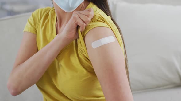 Midsection of asian woman in face mask sitting on sofa showing arm with plaster after vaccination alt