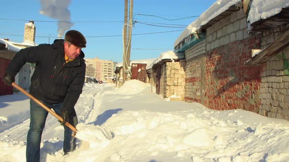 A Man in Warm Clothes with a Shovel Clears the Road of the Snow alt