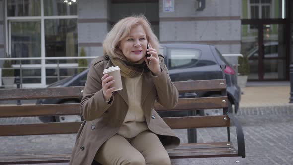 Portrait of Excited Senior Caucasian Woman Talking on the Phone As Sitting on Bench Outdoors alt