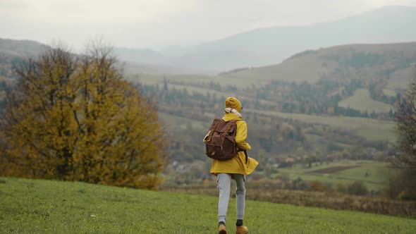 Slow Motion Happy Woman Hiker in Yellow Raincoat with Backpack Runs Away with Raised Arms in Green alt