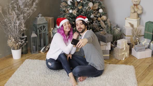 Wife and Husband in Santa's Hats Doing Selfie on Smartphone Near Christmas Tree.