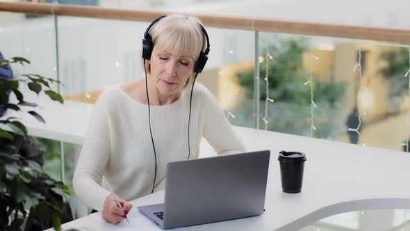 Mature Caucasian Woman Businesswoman Teacher in Headphones Looks at Laptop Screen Takes Notes Writes alt