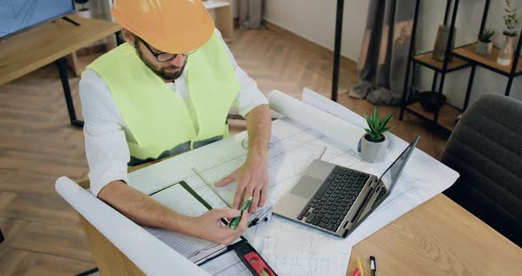 Designer in Helmet and Vest which Working with Architectural Scheme on Blueprint in Workroom alt