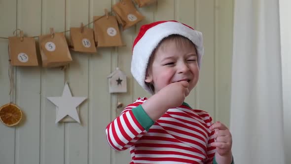Boy in Pajamas Santa Cap Eating Sweet Candy on Paper Advent Calendar with Presents Background alt