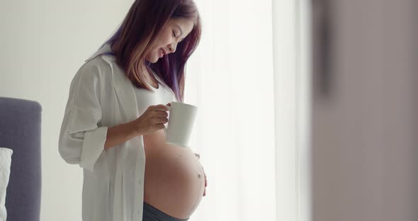 Young Asian Pregnant woman drinking coffee while standing beside a window in the home.