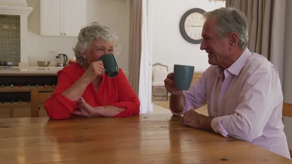 Happy caucasian senior couple sitting at table in dining room, smiling and drinking cups of coffee alt
