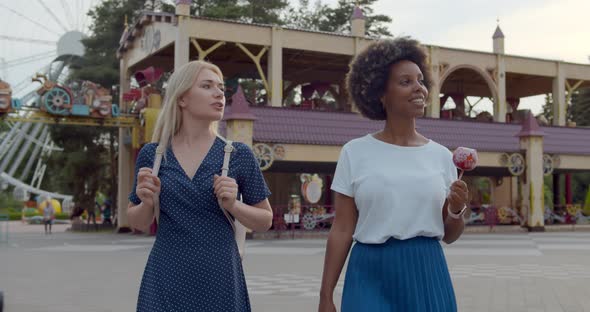 Happy Young Women Eating Candy Sweet and Walking at Amusement Park alt