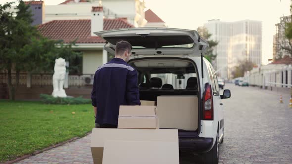 Young Handsome Smiling Caucasian Delivery Man with Boxes on Cart Moving Tovard to Van with Opened alt