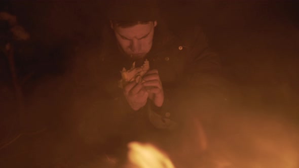 Homeless Man Eats Piece of Roll at Campfire in Night Forest alt