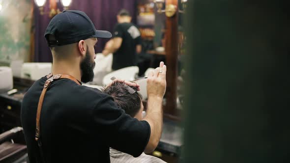 Side and Back View of the Stylish Barber with Beard Performing a Haircut with Scissors Holding a alt